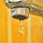 a close up shot of a water droplet hanging from a stainless steel faucet highlighting surface tensi